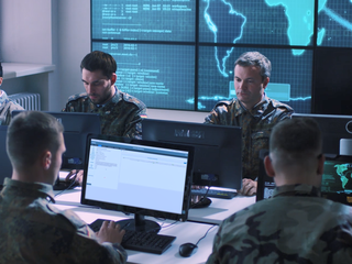 A group of soldiers is gathered indoors at an IMINT and GEOINT Centre, interacting with computers and discussing mission-related tasks. The setting includes intelligence displays and monitors, creating a professional work environment