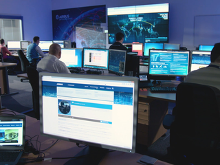 A group of people seated at computers in an indoor office environment at an IMINT and GEOINT Centre. 