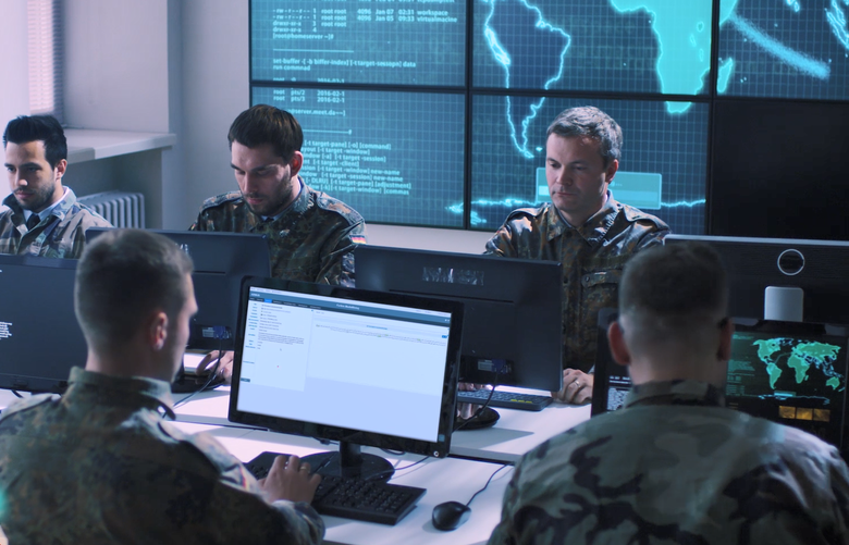 A group of soldiers is gathered indoors at an IMINT and GEOINT Centre, interacting with computers and discussing mission-related tasks. The setting includes intelligence displays and monitors, creating a professional work environment